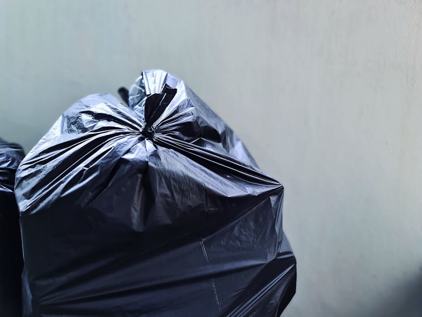 Close-up view of three black plastic rubbish bags, tied securely at the top, placed against a plain, light-colored wall. The bags appear to be made of shiny, smooth plastic material and are slightly crumpled with visible creases and folds. The largest bag is positioned in the foreground, occupying the left part of the image, with two smaller bags stacked behind it. The setting suggests an outdoor or semi-outdoor area, possibly a driveway or a designated refuse collection point, consistent with private waste disposal or rubbish removal services provided by Waste Collection Tooting. The background wall is neutral grey or off-white, with no additional environmental details visible, emphasizing the focus on the neatly tied rubbish bags ready for collection or disposal, underscoring the practical aspect of on-site waste management. The lighting appears natural or ambient, with no harsh shadows, highlighting the clean, organized state of the rubbish awaiting collection.