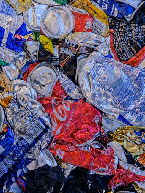 Close-up photograph of a heap of crushed aluminum beverage cans consisting of various brands, displaying metallic surfaces with textures ranging from smooth to crinkled. The cans are predominantly silver with some featuring red, blue, and black labels, including a partially visible Coca-Cola can in red. They are piled together haphazardly, creating a dense, chaotic arrangement with some cans overlapping others, and visible pull tabs and open tops. The background includes other crumpled cans and wrapped packaging, suggesting an indoor or controlled environment for waste collection. The lighting highlights the reflective qualities of the aluminum, emphasizing the metallic sheen and varying shades of grey. This scene depicts an example of recyclable waste possibly collected for disposal or recycling services by Waste Collection Tooting, illustrating the process of managing household or commercial rubbish through private waste handling methods, aligning with alternative rubbish collection services in the area.