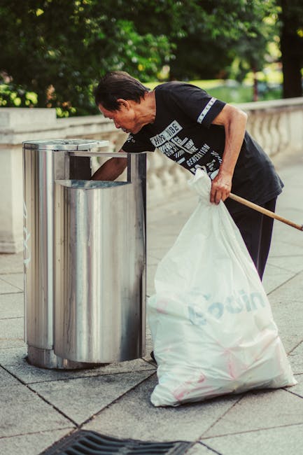 A man wearing a black t-shirt is bending forward as he disposes of a white plastic bag filled with rubbish into a stainless steel recycling bin situated on a paved sidewalk. The trash bin, part of a pair of modern, cylindrical containers, has a polished, reflective surface and is positioned near a low stone wall with a decorative balustrade. The background includes lush green trees and a grassy area, suggesting an outdoor public space. The man’s arm is extended, with his hand grasping the bag, while he holds a wooden stick in his other hand, possibly for cleaning or supporting the waste collection process. This scene relates to the provision of private rubbish collection services in the area, exemplifying on-site waste disposal efforts that align with independent waste management practices aimed at maintaining cleanliness in public parks or communal outdoor areas. Waste Collection Tooting, a professional waste management service, facilitates such rubbish removal activities, supporting residents and visitors in maintaining tidy environments.