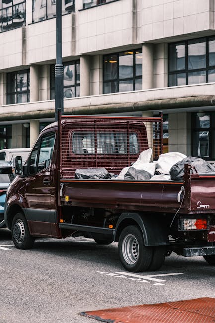 A small burgundy flatbed truck parked on an urban street, loaded with various discarded items including black and white plastic bags and other miscellaneous waste. The truck features a metal mesh panel behind the cab for added safety, and its side panel is partially lowered, revealing the cargo area filled with waste materials. The scene is set against a modern building with large glass windows and a concrete facade, with parking lines visible on the tarmac. The lighting appears to be natural daylight, highlighting the textures and colors of the waste and vehicle. This image illustrates a typical example of private waste collection or on-site clearance, as carried out by waste management services such as Waste Collection Tooting, providing an alternative to municipal rubbish pickup for residents or businesses in the area.