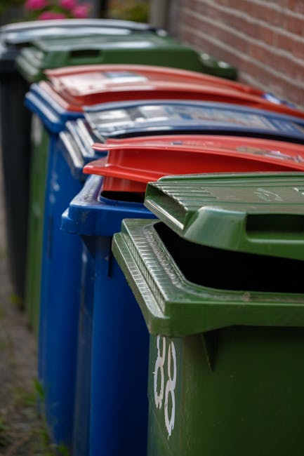 A row of large outdoor waste bins is positioned along a pavement adjacent to a brick wall, with each bin exhibiting a distinct color and material. The closest bin in the foreground is green with a smooth, matte finish, featuring a closed lid and a slightly ribbed surface pattern. Behind it, a blue bin with a similar matte texture is visible, with its lid partially open, revealing an empty interior. Further back, a red bin with a glossy finish and curved lid is stacked neatly on top of a blue bin, with its lid slightly ajar. In the background, a dark green bin with a matte surface and a partially visible lid is positioned alongside other bins, all aligned closely in a straight row along the edge of a sidewalk. The environment suggests an organized collection point, supported by Waste Collection Tooting’s services, for private refuse disposal or alternative waste handling outside residential or commercial premises. The scene is evenly lit, highlighting the clean, maintained condition of the bins and their surrounding area, with no signs of waste overflowing or environmental clutter.