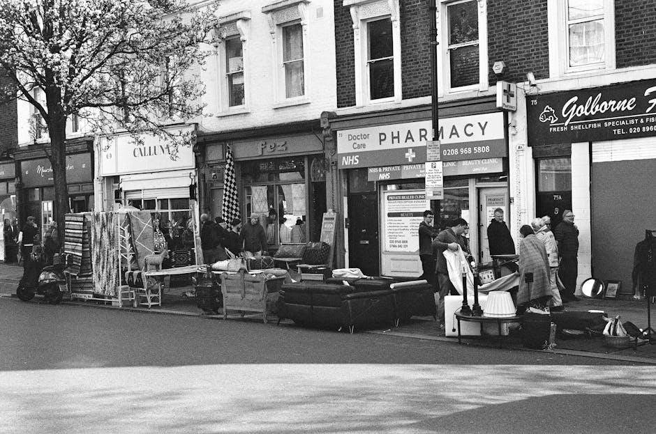 A street scene featuring a variety of second-hand furniture and household items displayed on the pavement outside storefronts, including a sofa, armchair, and several tables, which are arranged in front of shops, possibly indicating a street sale or cleanup event. The furniture surfaces appear to be upholstered, with some items showing signs of use, and the furniture is placed directly on the pavement beneath a row of tall brick buildings with large windows. Several pedestrians are gathered around the items, some engaging in conversation, while others browse or pause to view the displayed goods. In the background, commercial signs indicate businesses such as a pharmacy and a fish shop, with a few trees and street signs also visible. The scene is illuminated by natural daylight, capturing an ordinary, busy urban environment, which aligns with private or independent rubbish clearance services provided by Waste Collection Tooting, focusing on the removal of bulky items from residential or commercial premises.