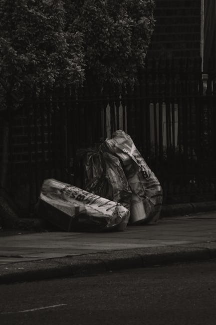 Three large black rubbish bags made of glossy plastic material are placed on a pavement curb, with some of the bags appearing crumpled and slightly torn, revealing the compressed waste inside. The bags are positioned close together, leaning slightly to the right, with their openings visible on the upper side. Behind them, a dark metal fence with vertical bars and a dense canopy of leafy branches extends into the background, casting shadows on the scene. The surrounding environment suggests an urban street setting, with a section of asphalt road in the foreground and faint hints of a building structure or garden area behind the fence. The lighting appears to be subdued, possibly during the late evening or overcast day, emphasizing the reflective surface of the rubbish bags. This scene is indicative of private waste disposal or independent rubbish removal services, consistent with non-public collection methods. Waste Collection Tooting’s focus on rubbish services aligns with the typical removal of bulky waste or refuse in urban residential areas, as depicted in this image.